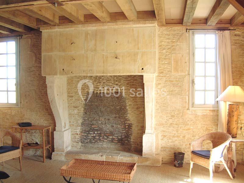 Cheminée en pierre ancienne dans une pièce lumineuse avec murs en pierre, poutres apparentes et mobilier en osier.