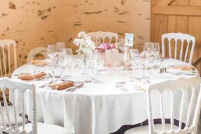 Salle en pierre avec plafond en poutres apparentes, tables dressées pour un repas, chaises blanches alignées.