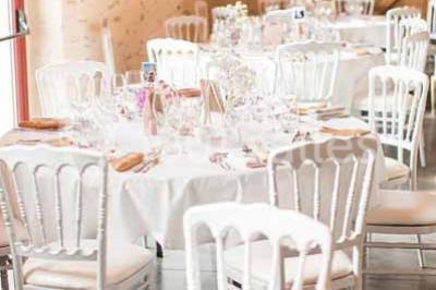 Salle en pierre avec plafond en poutres apparentes, tables dressées pour un repas, chaises blanches alignées.