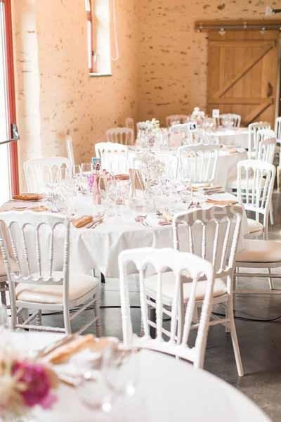 Salle décorée pour un événement avec des tables rondes dressées, nappes blanches et chaises blanches élégantes.