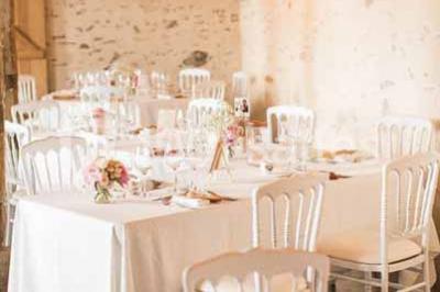 Salle en pierre avec plafond en poutres apparentes, tables dressées pour un repas, chaises blanches alignées.