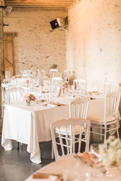 Salle de réception lumineuse avec tables dressées, nappes blanches, chaises en bois et décorations florales.