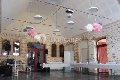 Salle en pierre avec plafond en poutres apparentes, tables dressées pour un repas, chaises blanches alignées.