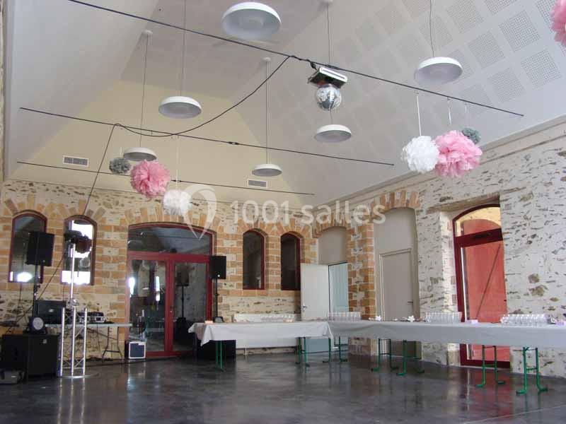 Salle en pierre avec tables alignées, décorée de pompons suspendus roses et blancs, éclairée par des luminaires modernes.