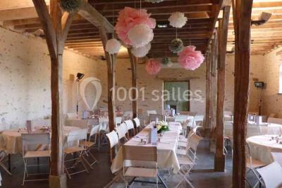 Salle en pierre avec plafond en poutres apparentes, tables dressées pour un repas, chaises blanches alignées.