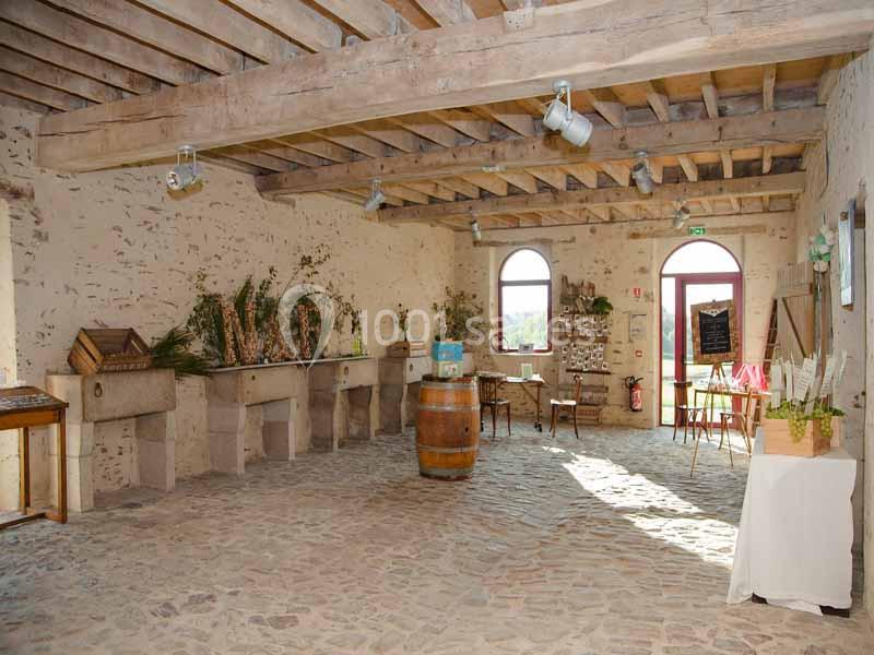 Salle en pierre avec poutres apparentes, décorée de fleurs et de plantes, éclairée par des fenêtres en arche.