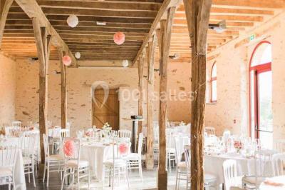 Salle en pierre avec plafond en poutres apparentes, tables dressées pour un repas, chaises blanches alignées.