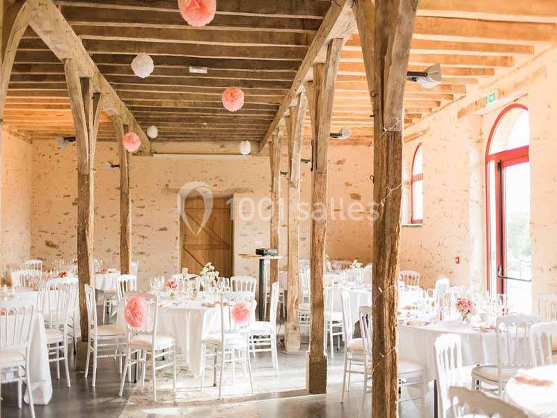 Salle de réception rustique avec poutres apparentes, tables rondes dressées et décorations florales et suspendues.