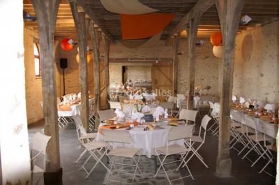 Salle en pierre avec plafond en poutres apparentes, tables dressées pour un repas, chaises blanches alignées.