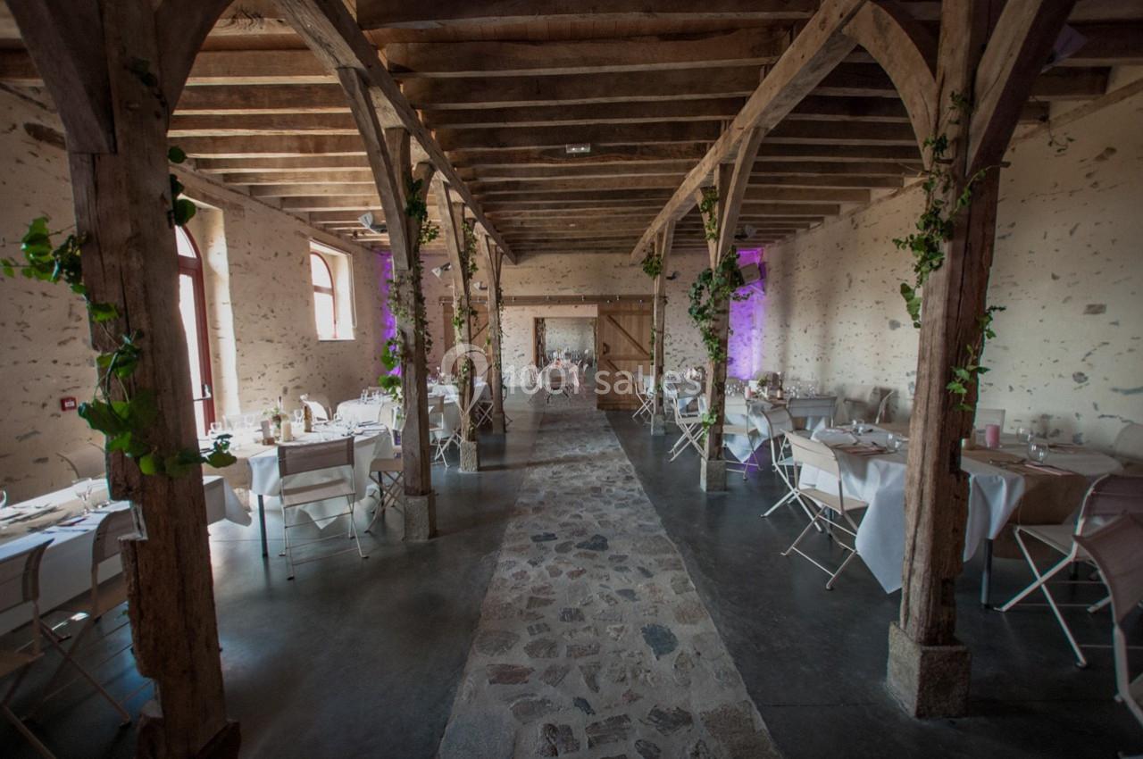 Salle rustique avec poutres en bois, tables dressées et décorations végétales, éclairée par des fenêtres et des lumières…