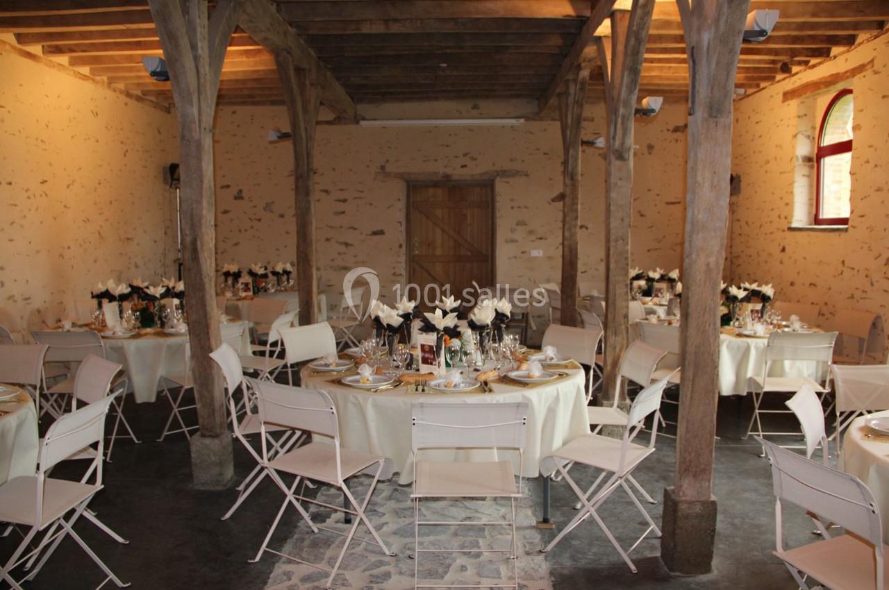 Salle rustique aménagée pour un repas, avec tables rondes dressées, chaises blanches pliantes et décorations florales.