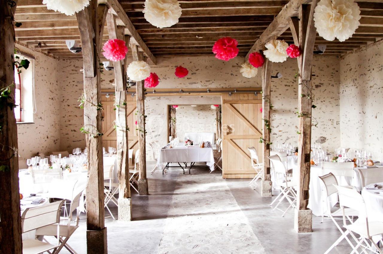 Salle de réception rustique décorée de pompons suspendus rouges et blancs, avec tables dressées et chaises blanches.