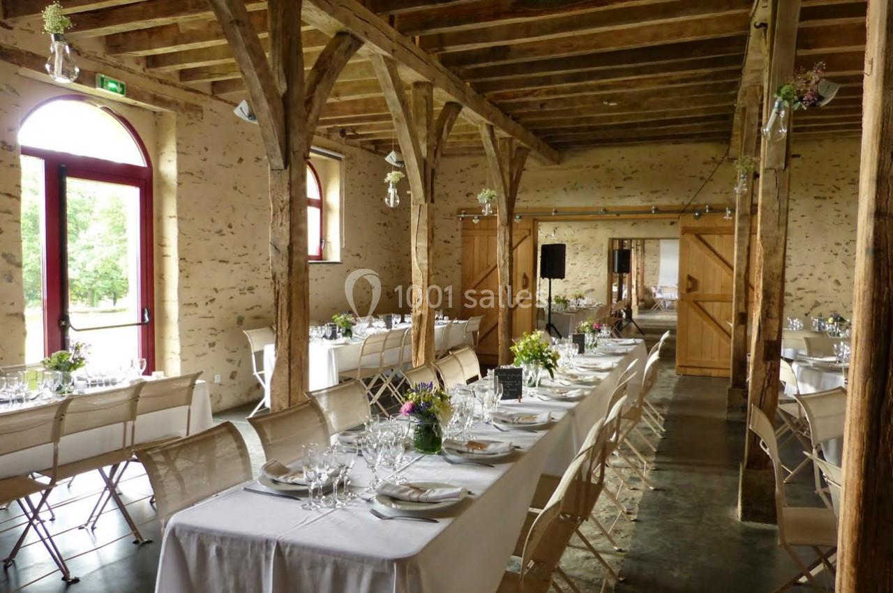 Salle de réception rustique avec poutres apparentes, tables dressées avec nappes blanches et décoration florale.