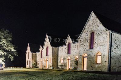 Salle en pierre avec plafond en poutres apparentes, tables dressées pour un repas, chaises blanches alignées.
