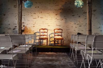 Salle en pierre avec plafond en poutres apparentes, tables dressées pour un repas, chaises blanches alignées.