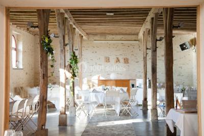 Salle en pierre avec plafond en poutres apparentes, tables dressées pour un repas, chaises blanches alignées.