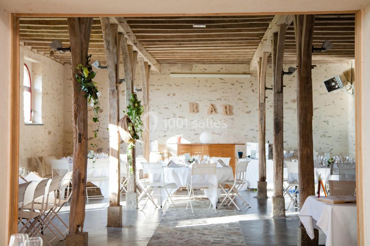 Salle de réception lumineuse avec poutres apparentes, tables dressées et espace bar en arrière-plan.