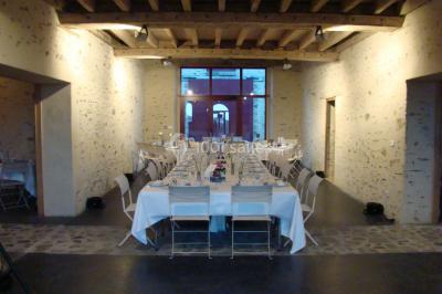 Salle en pierre avec plafond en poutres apparentes, tables dressées pour un repas, chaises blanches alignées.