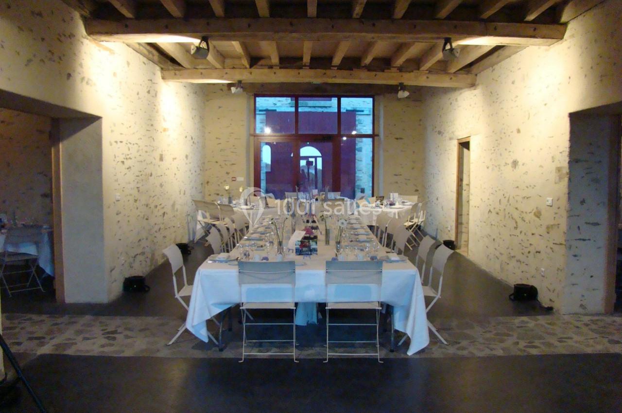 Salle en pierre avec plafond en poutres apparentes, tables dressées pour un repas, chaises blanches alignées.