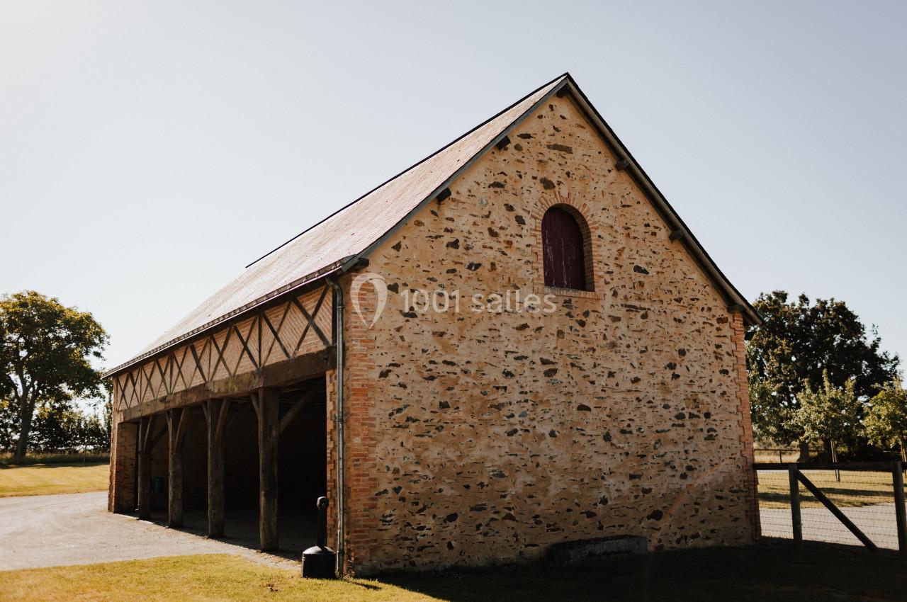 Bâtiment en pierre avec toit en pente, ouvertures en bois et petite fenêtre en arc, situé dans un espace verdoyant.