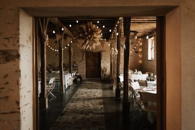 Salle en pierre avec plafond en poutres apparentes, tables dressées pour un repas, chaises blanches alignées.
