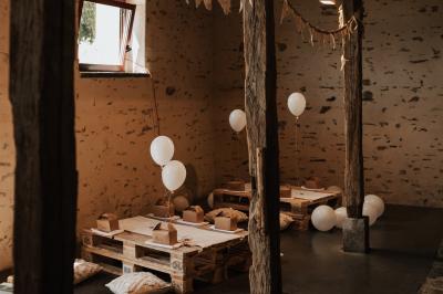 Salle en pierre avec plafond en poutres apparentes, tables dressées pour un repas, chaises blanches alignées.