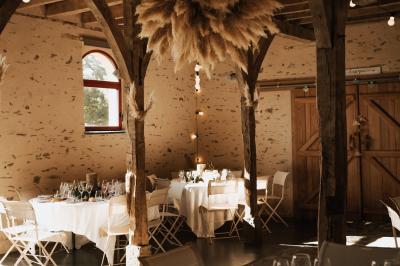 Salle en pierre avec plafond en poutres apparentes, tables dressées pour un repas, chaises blanches alignées.
