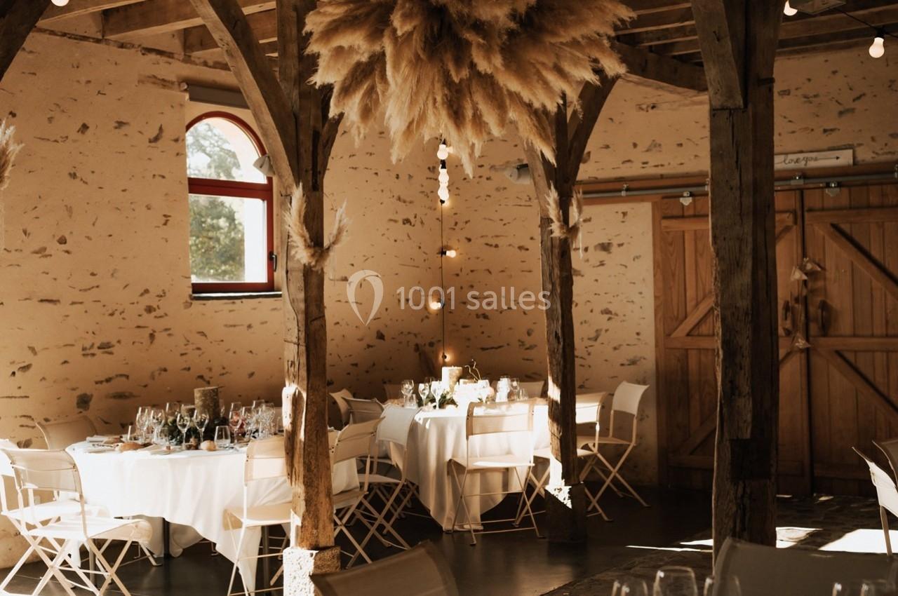 Salle de réception rustique avec tables rondes dressées, chaises pliantes blanches et décorations en pampas suspendues.