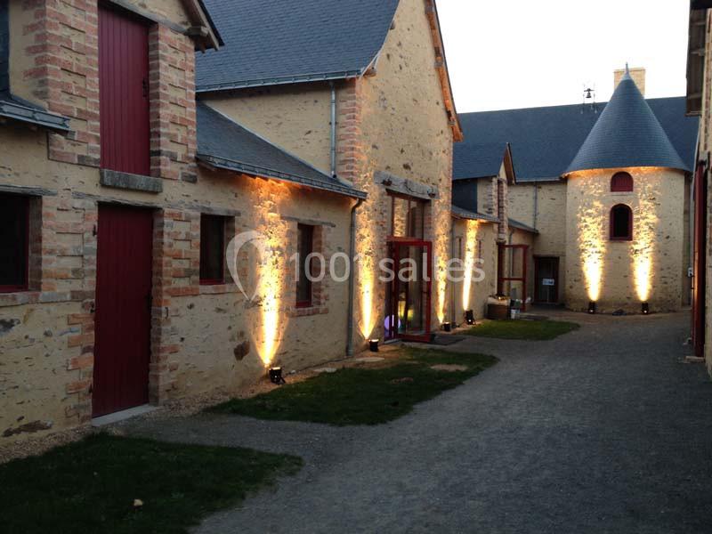 Façade en pierre éclairée par des lumières au sol, avec portes et fenêtres rouges, dans une cour pavée.