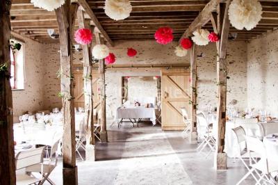 Salle en pierre avec plafond en poutres apparentes, tables dressées pour un repas, chaises blanches alignées.