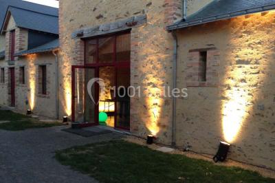 Salle en pierre avec plafond en poutres apparentes, tables dressées pour un repas, chaises blanches alignées.