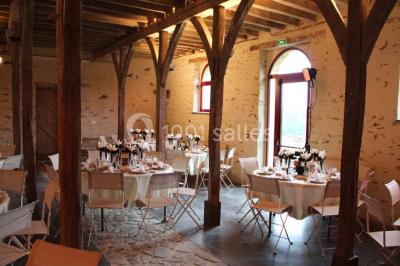 Salle en pierre avec plafond en poutres apparentes, tables dressées pour un repas, chaises blanches alignées.