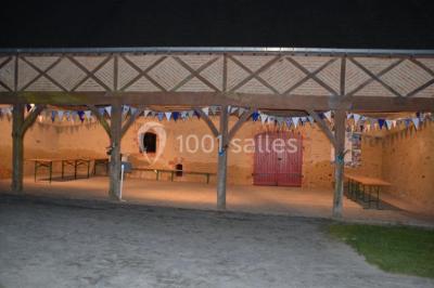Salle en pierre avec plafond en poutres apparentes, tables dressées pour un repas, chaises blanches alignées.