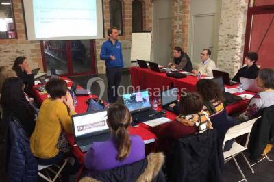 Salle de réception rustique avec tables rondes et rectangulaires dressées pour un événement, éclairage tamisé.