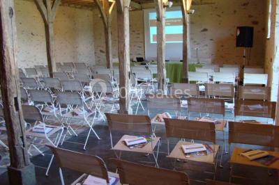 Salle de réception rustique avec tables rondes et rectangulaires dressées pour un événement, éclairage tamisé.