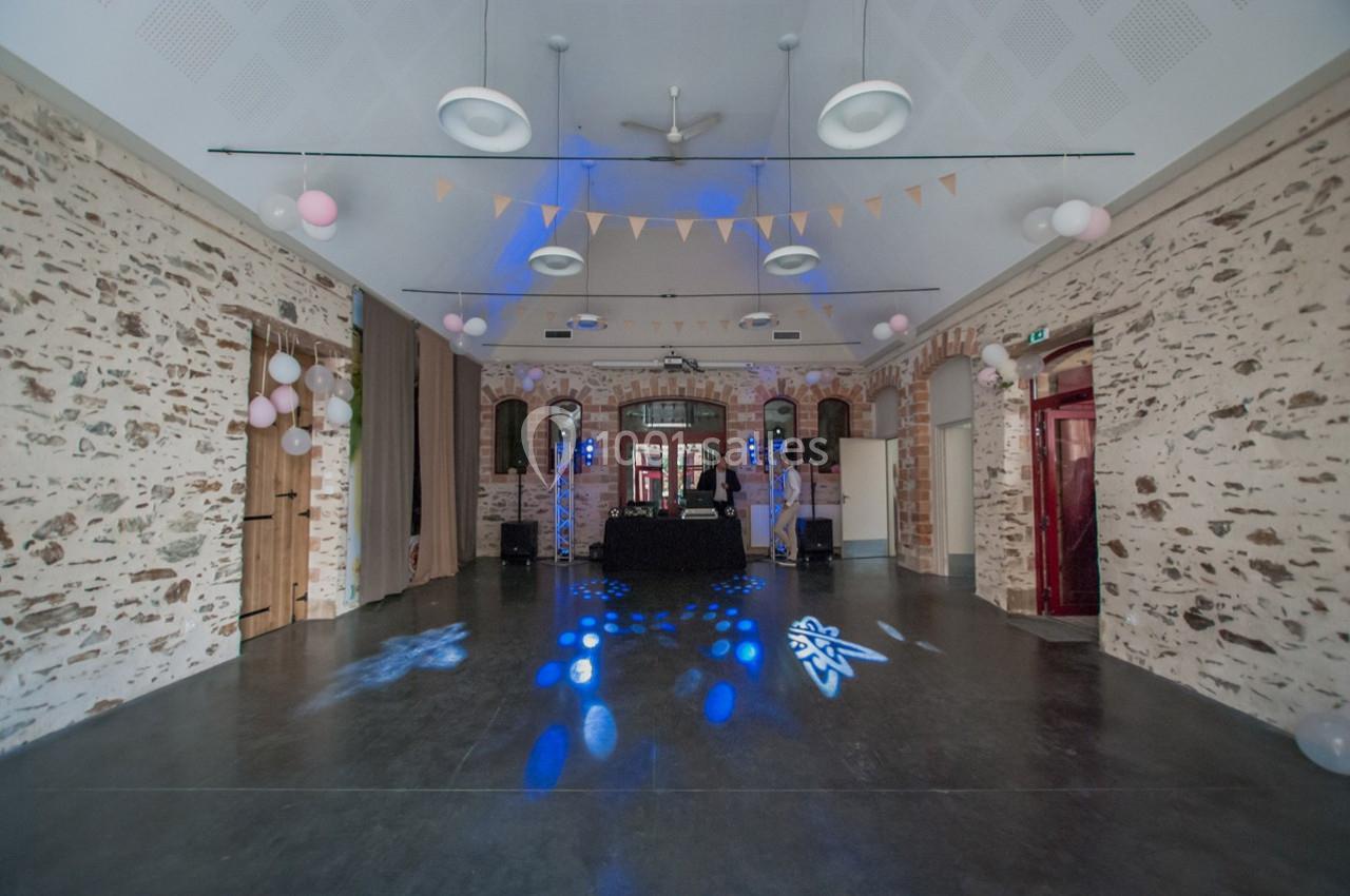 Salle en pierre décorée de guirlandes et ballons, avec une piste de danse éclairée et une table de DJ au centre.