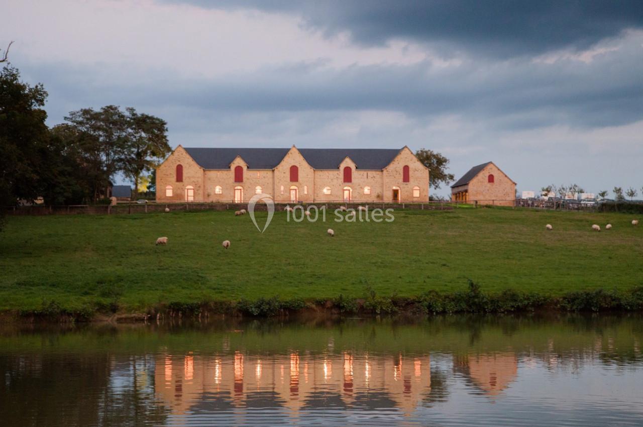 Bâtiments en pierre avec fenêtres rouges, situés sur une colline verdoyante près d'un étang reflétant leur image.