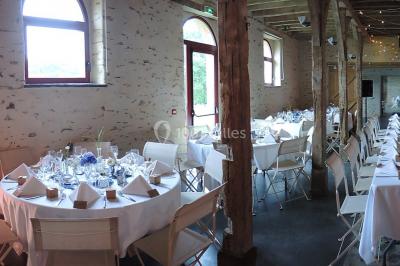 Salle de réception rustique avec tables rondes et rectangulaires dressées pour un événement, éclairage tamisé.