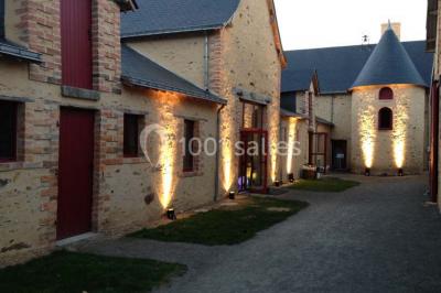 Salle de réception rustique avec tables rondes et rectangulaires dressées pour un événement, éclairage tamisé.