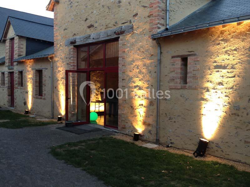 Façade en pierre d'un bâtiment éclairée par des projecteurs au sol, avec une porte vitrée ouverte.