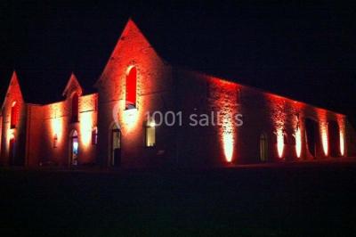 Salle de réception rustique avec tables rondes et rectangulaires dressées pour un événement, éclairage tamisé.