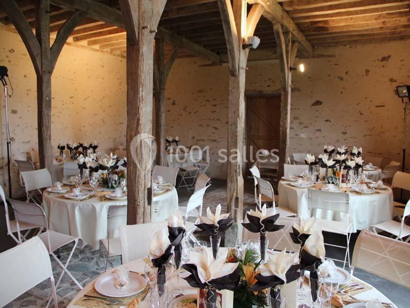 Salle rustique avec poutres apparentes, tables rondes décorées de nappes blanches et centres de table floraux.