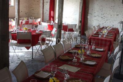 Salle de réception rustique avec tables rondes et rectangulaires dressées pour un événement, éclairage tamisé.