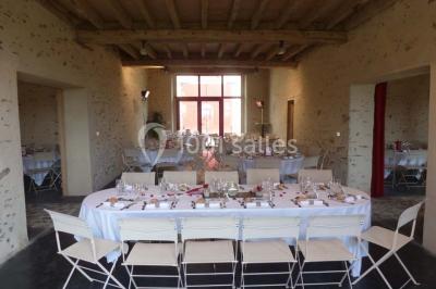 Salle de réception rustique avec tables rondes et rectangulaires dressées pour un événement, éclairage tamisé.