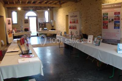 Salle de réception rustique avec tables rondes et rectangulaires dressées pour un événement, éclairage tamisé.