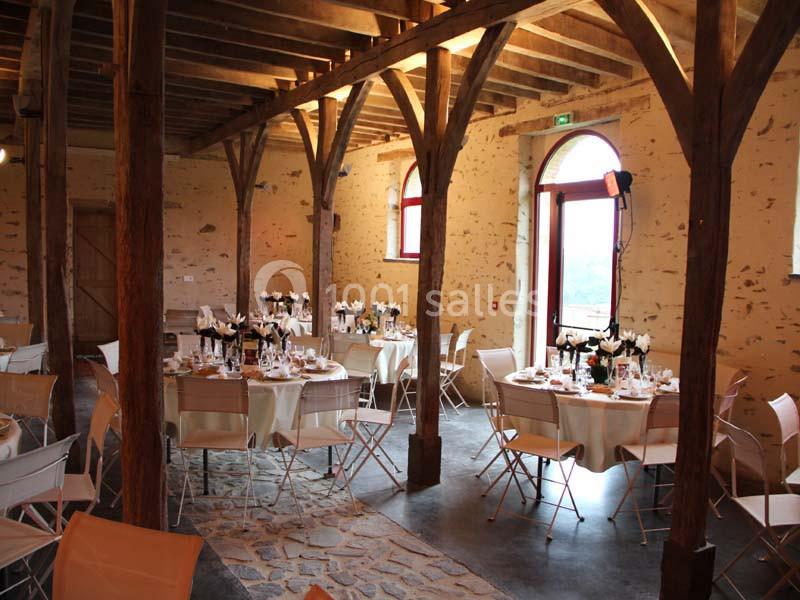 Salle de réception rustique avec tables dressées, chaises pliantes et poutres en bois apparentes.