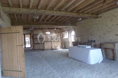 Salle de réception rustique avec tables rondes et rectangulaires dressées pour un événement, éclairage tamisé.