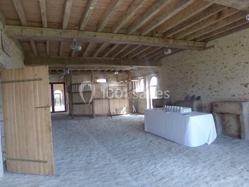 Salle en pierre avec poutres apparentes, sol en sable, table couverte d'une nappe blanche et portes en bois ouvertes.