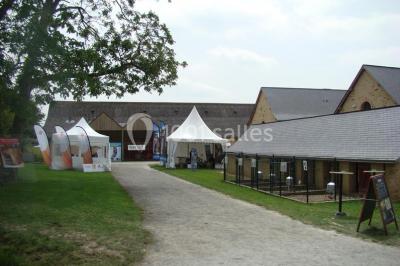 Salle de réception rustique avec tables rondes et rectangulaires dressées pour un événement, éclairage tamisé.