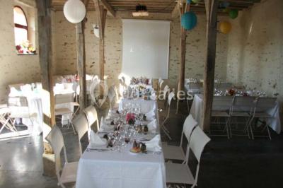 Salle de réception rustique avec tables rondes et rectangulaires dressées pour un événement, éclairage tamisé.